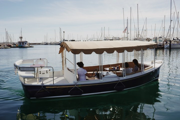 A small covered boat in a marina with three people onboard, surrounded by many docked sailboats.